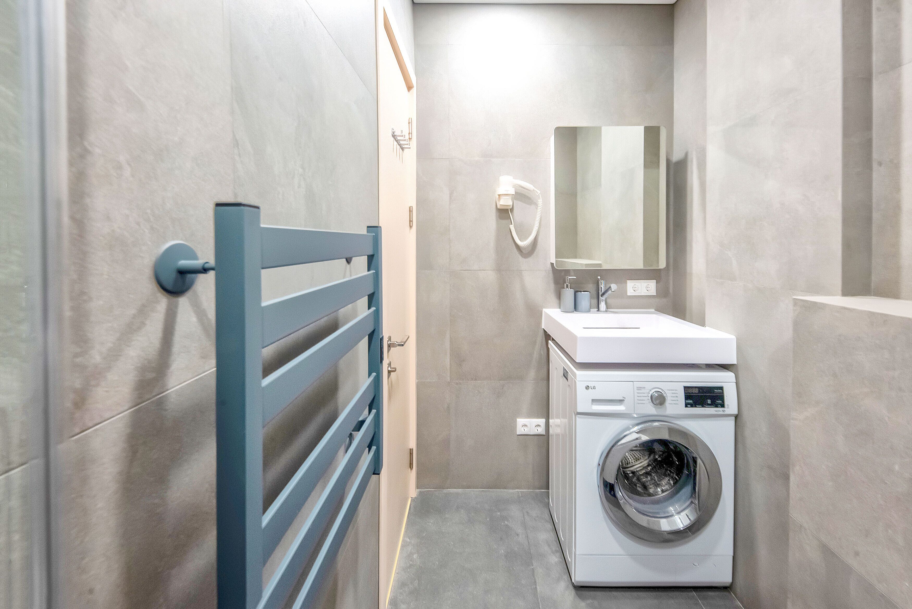 Une petite salle de bain avec un mur recouvert de carreaux gris, un lave-linge blanc, un lavabo avec un miroir et un porte-serviettes bleu.