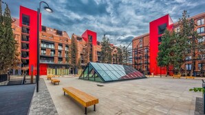 Ein moderner Stadtplatz mit einem pavilionsartigen Bauwerk unter Glasdach, Bänken und roten Gebäuden.