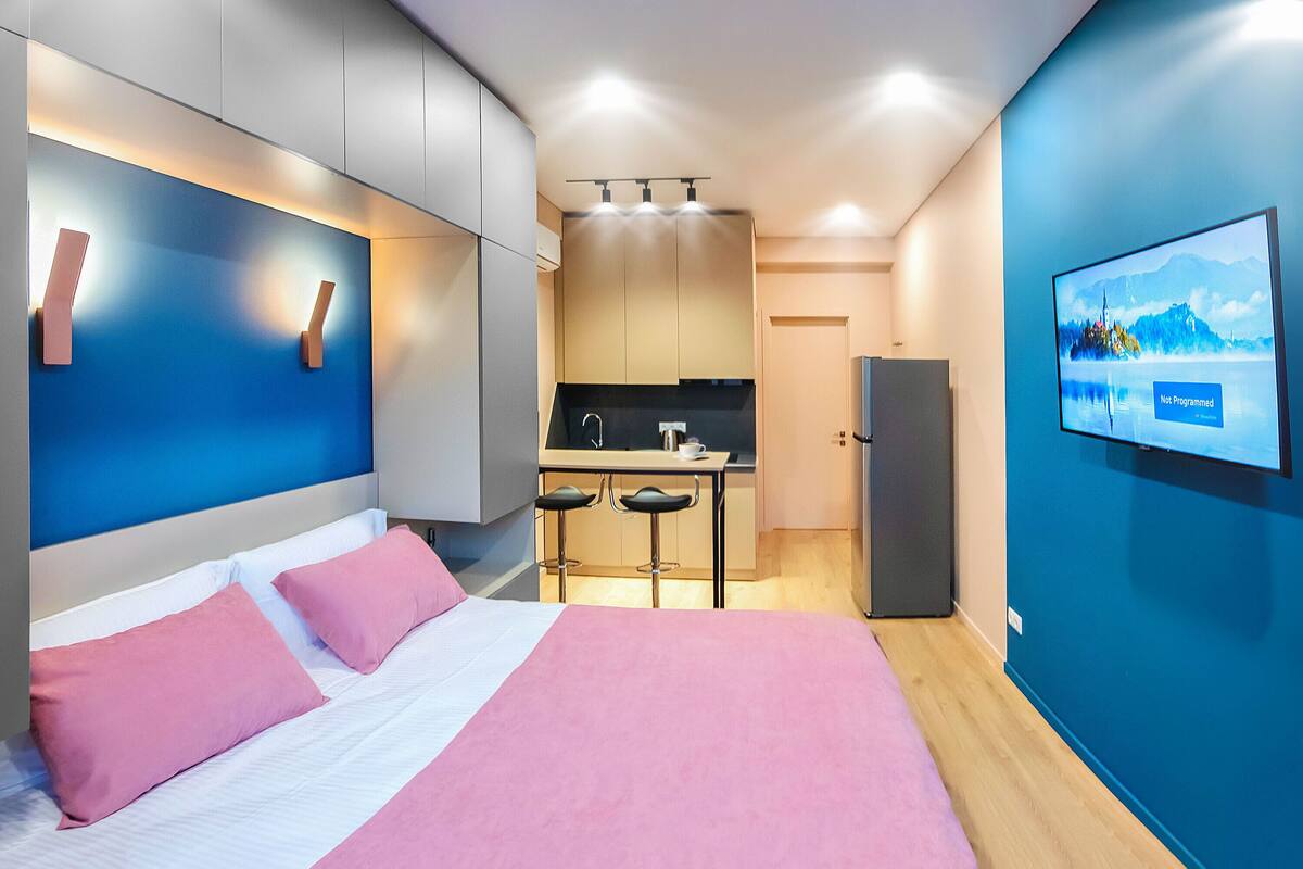 A modern hotel room with a bed, a small kitchenette, and a TV displaying a scenic landscape.