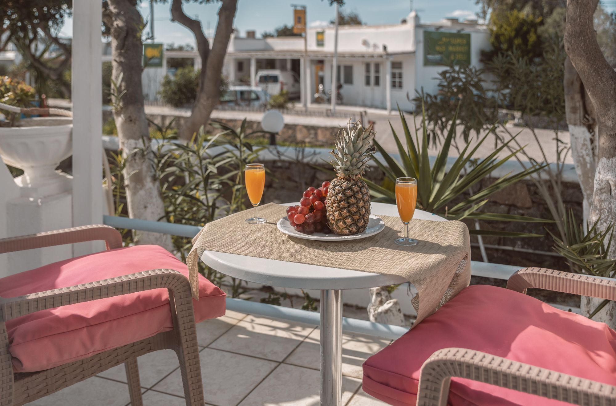 A table with a plate of fruit and two glasses of orange juice, set on a balcony with wicker chairs and a view of buildings and trees.