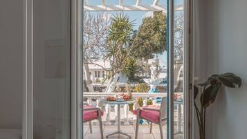 Vista a través de una puerta de vidrio a un patio con una pérgola blanca, mobiliario exterior y diversas plantas.