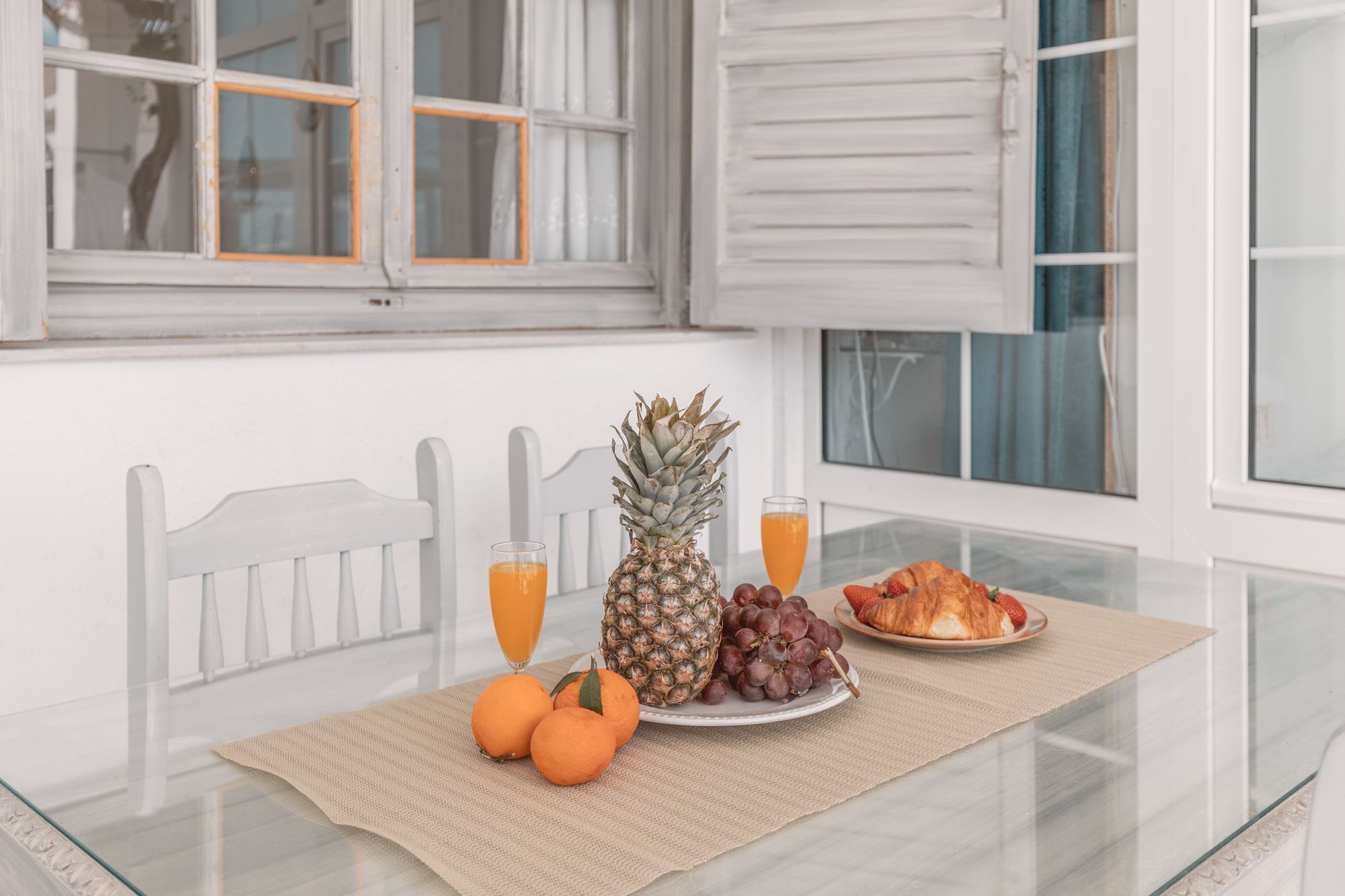 A breakfast setting with a pineapple, grapes, oranges, a croissant, and orange juice on a glass table with a white chair.