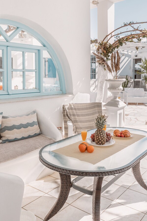 Un patio iluminado por el sol con una mesa de vidrio, muebles blancos y vista al mar a través de una ventana con marco azul.