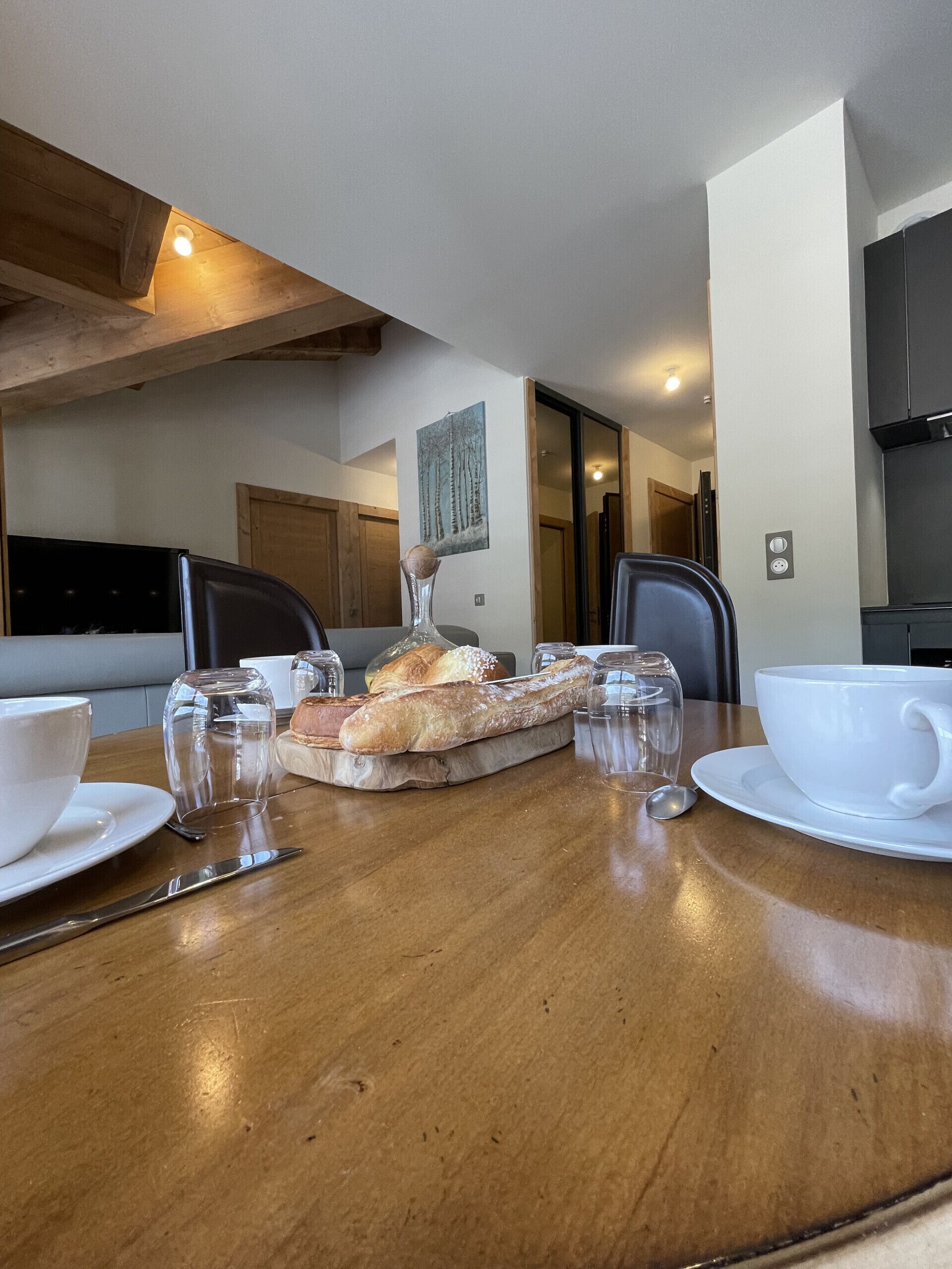 Una mesa de comedor con pan, una taza blanca y un vaso. Hay sillas alrededor de la mesa y un techo de madera.