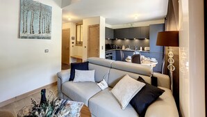 Una sala de estar moderna con un sofá, una mesa de centro y un comedor con mesa y sillas. Se observa una cocina al fondo con electrodomésticos modernos.