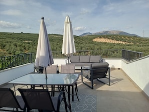 Un área de estar al aire libre con mesa y sillas, dos sombrillas y vistas a un paisaje montañoso.