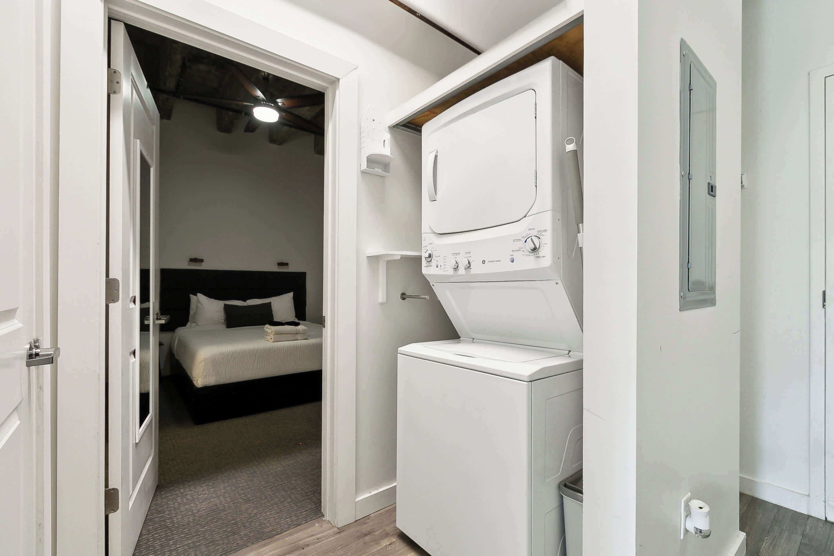 A compact room with a bed, a washing machine, and a door leading to another room.