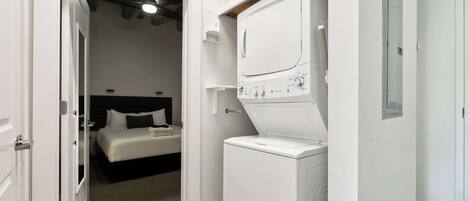 A compact room with a bed, a washing machine, and a door leading to another room.