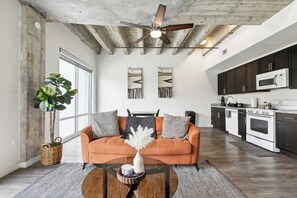 Un salon moderne avec un canapé, une table basse et des coussins décoratifs. On aperçoit au fond un ventilateur de plafond et une cuisine équipée.