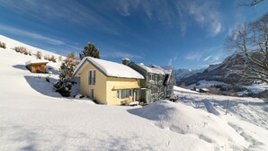 Uma casa de dois andares com telhado coberto de neve, rodeada por uma paisagem nevada e montanhas ao fundo.