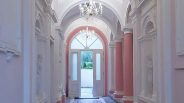 Un pasillo con paredes rosas, columnas blancas y una alfombra estampada que conduce a un amplio arco.