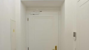 Un pasillo estrecho con una puerta blanca, un interruptor de luz y un pequeño cuadro eléctrico en la pared.