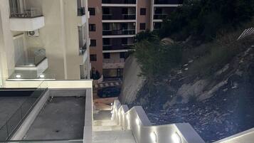 Ein mehrstöckiges Gebäude mit Balkonen, einer Treppe, die zu einer unteren Ebene führt, und Blick auf einen felsigen Hügel.