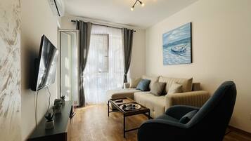 Ein modernes Wohnzimmer mit Sofa, Sessel und Couchtisch. Ein Flachbildfernseher ist an der Wand montiert, und ein Gemälde eines Bootes hängt über dem Sofa.