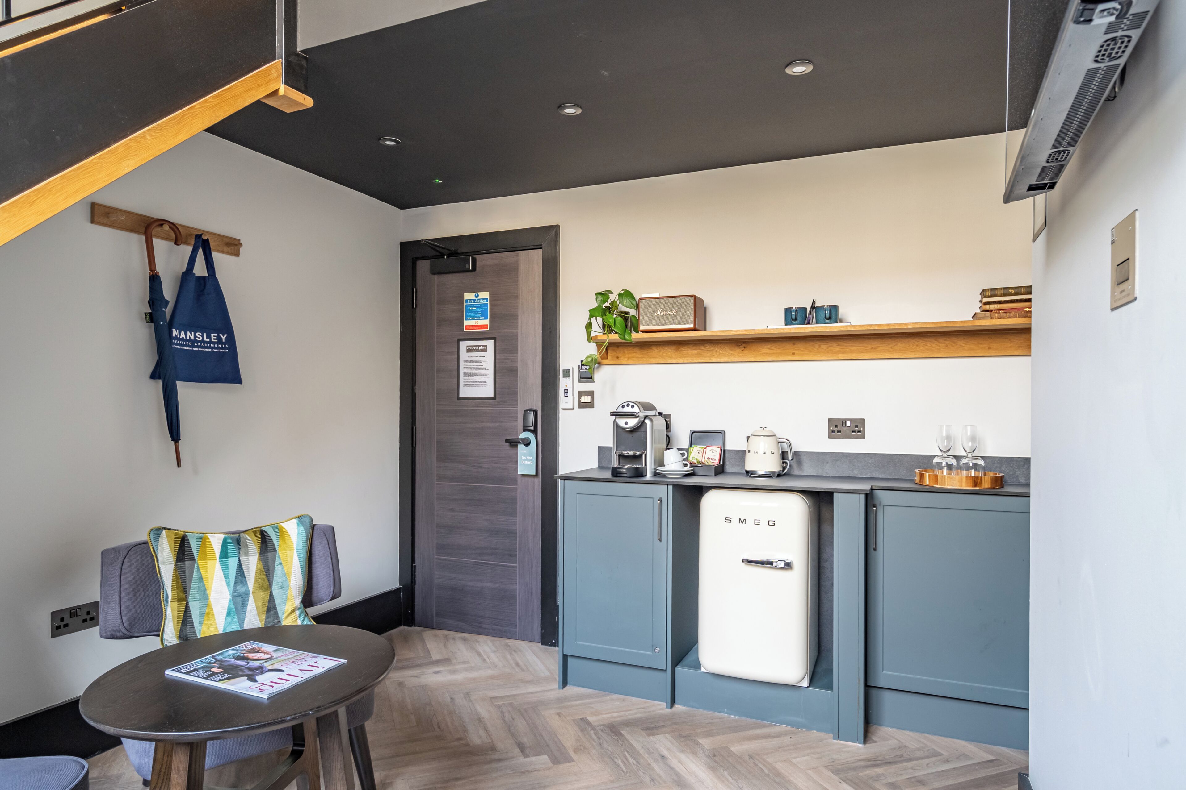 A compact kitchen with a refrigerator, coffee machine, and a shelf with cups and a plant.