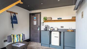 A compact kitchen with a refrigerator, coffee machine, and a shelf with cups and a plant.