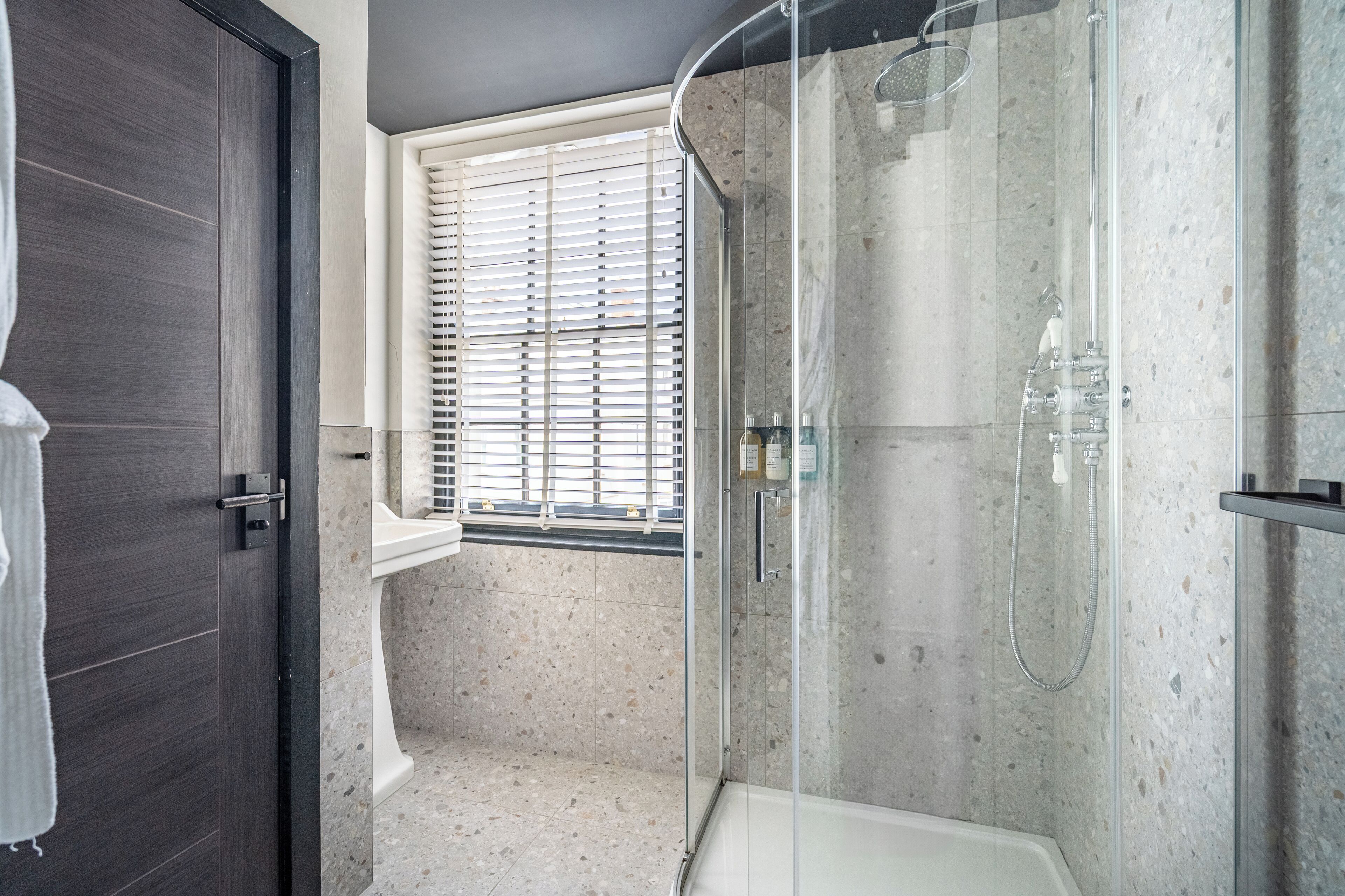 Une salle de bain moderne avec une douche en verre, une fenêtre avec des stores et une porte en bois foncé.