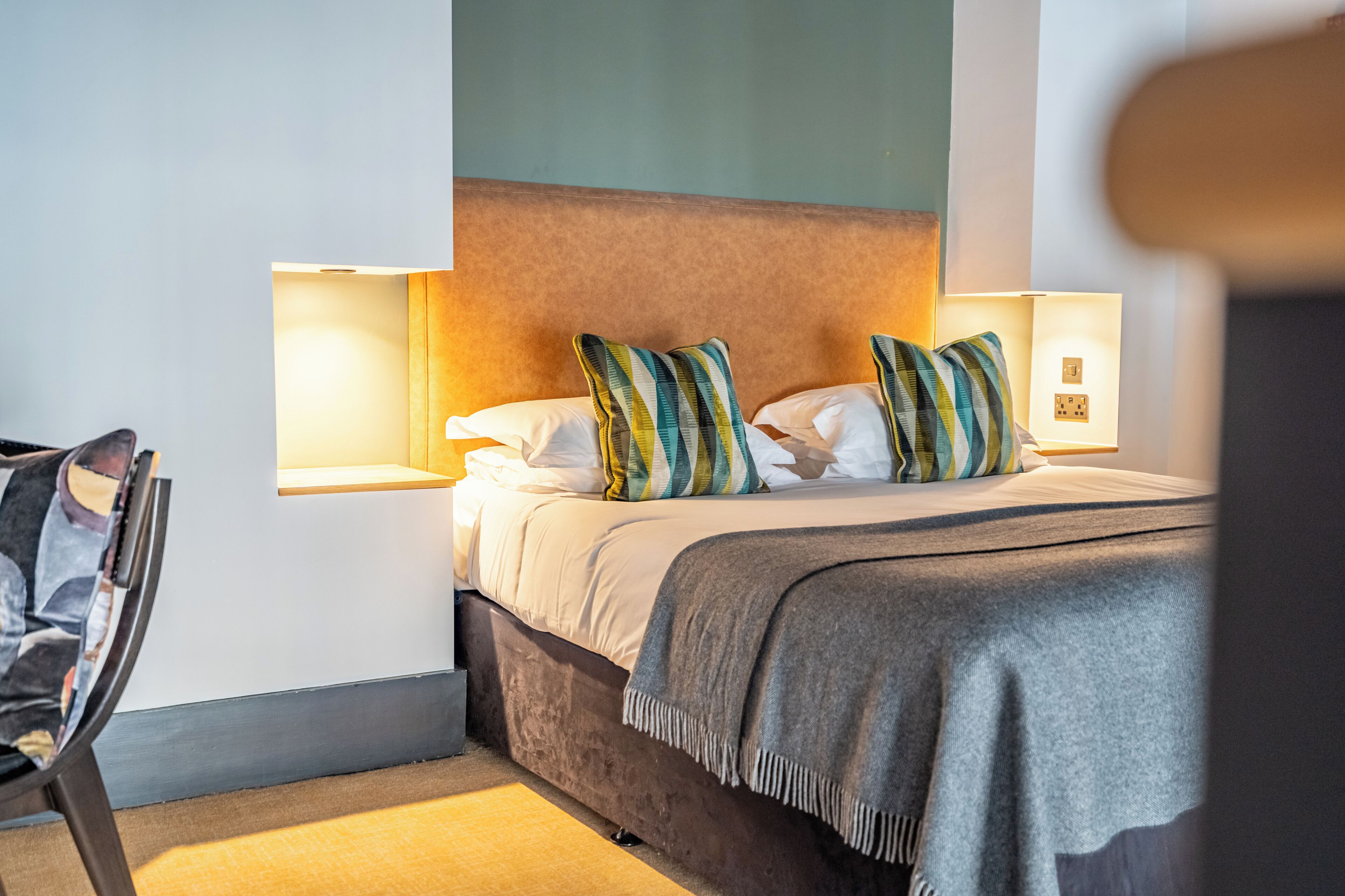 A neatly made bed with pillows, a bedside lamp, and a chair in a hotel room.