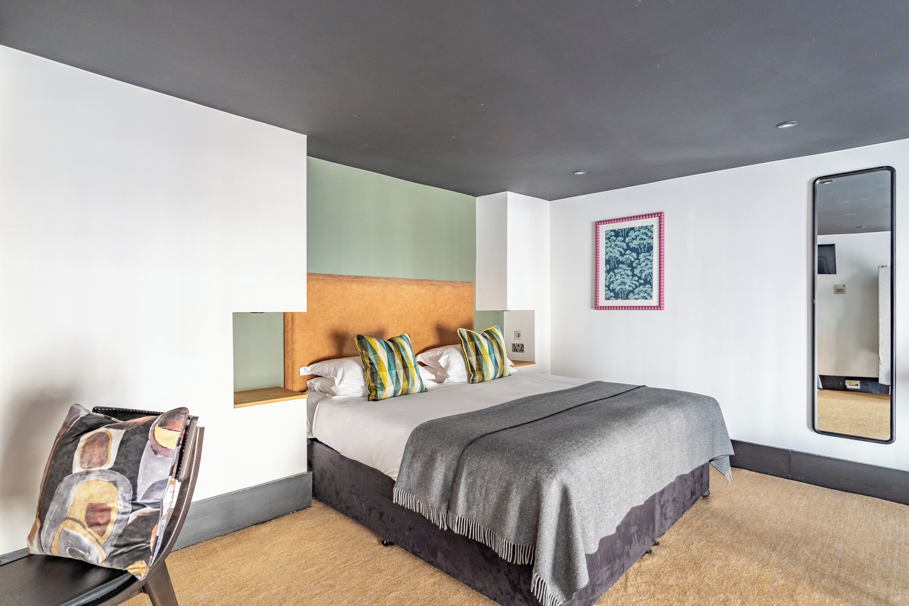 A modern hotel room with a large bed, a headboard, a bedside table, a chair, a mirror, and a framed artwork on the wall.