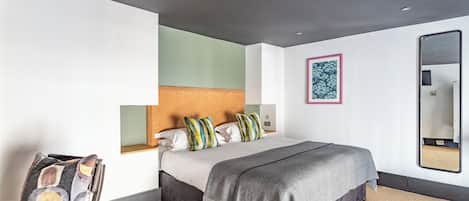 A modern hotel room with a large bed, a headboard, a bedside table, a chair, a mirror, and a framed artwork on the wall.