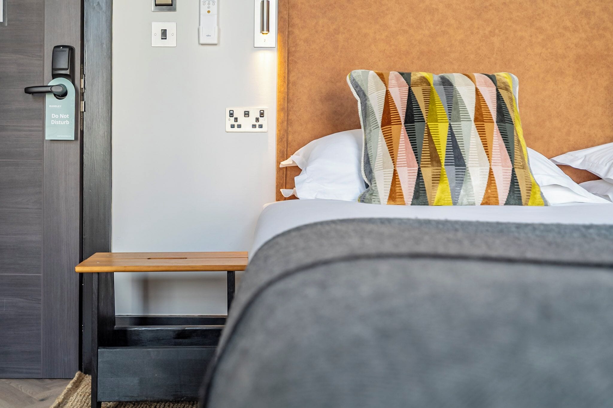 Ein Hotelzimmer mit Bett, Nachttisch, einer Tür mit Schloss und einer wandmontierten Bedienfeld.