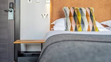 Ein Hotelzimmer mit Bett, Nachttisch, einer Tür mit Schloss und einer wandmontierten Bedienfeld.