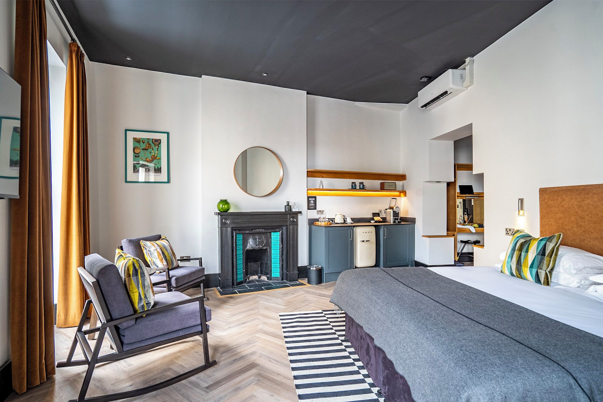 Ett modernt hotellrum med en säng, en sittgrupp med stolar, en öppen spis, ett litet kök och ett stort fönster med gardiner.