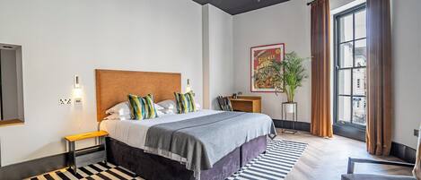 Un dormitorio moderno con una cama grande, una mesita de noche pequeña, una planta en maceta y una ventana con cortinas.