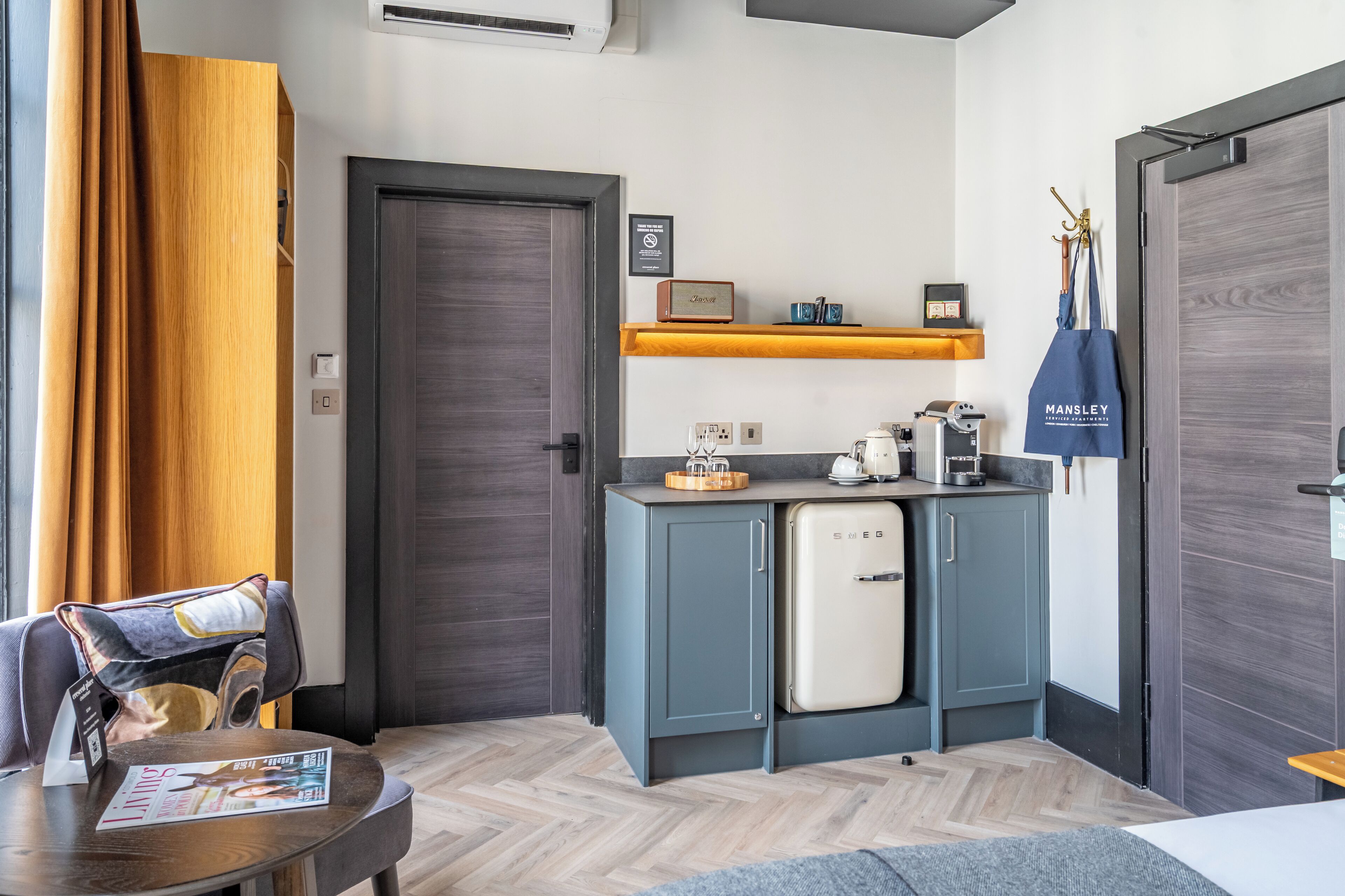 Cocina compacta con refrigerador, microondas y cafetera. Hay un estante de madera sobre la encimera con una jarra de café y un altavoz pequeño.