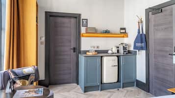 Cocina compacta con refrigerador, microondas y cafetera. Hay un estante de madera sobre la encimera con una jarra de café y un altavoz pequeño.