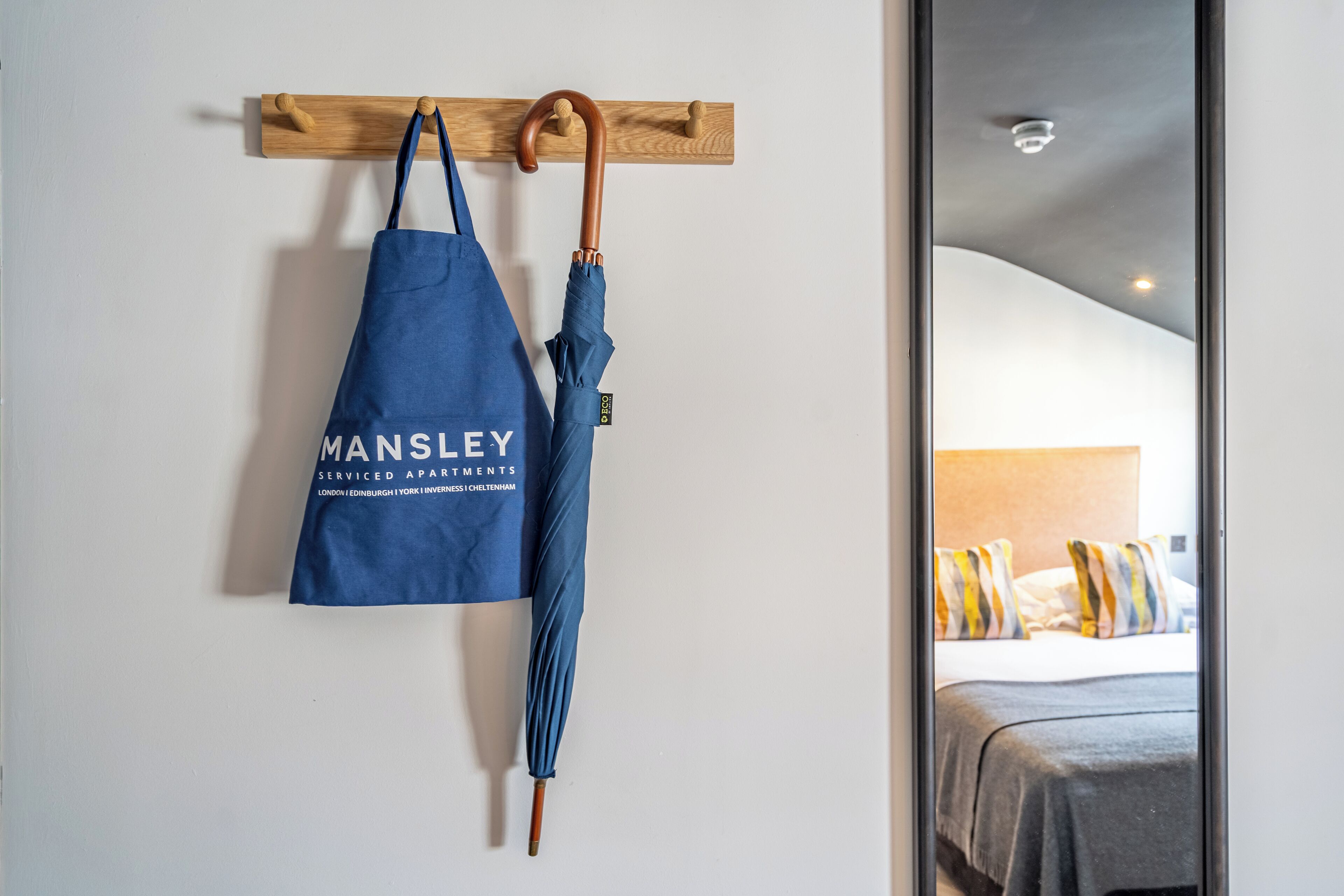 A blue bag with 'MANSLEY' text hanging on a wooden wall-mounted rack, next to a wooden-handled umbrella.