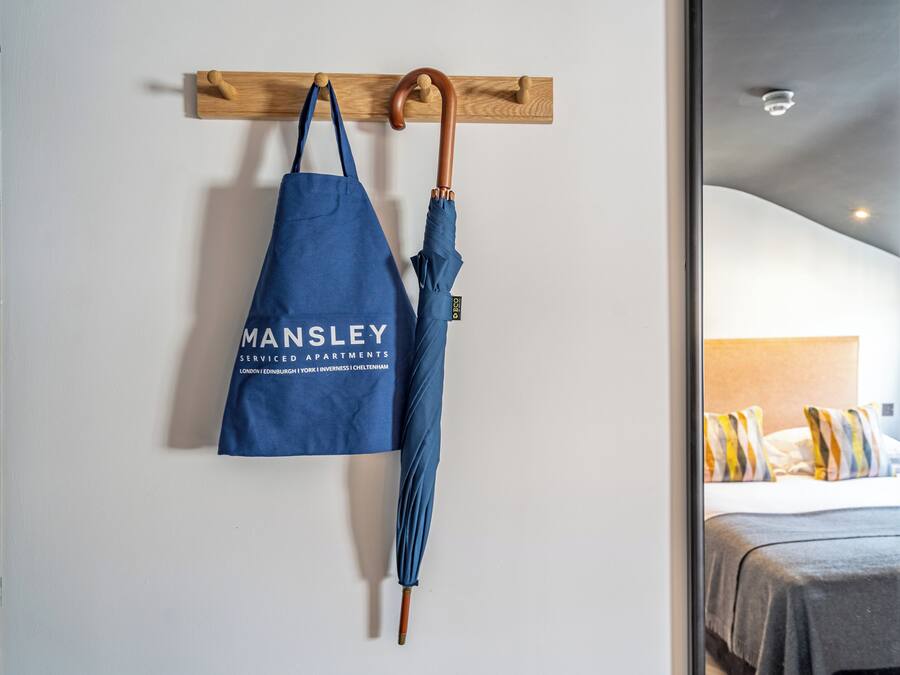 A blue bag with 'MANSLEY' text hanging on a wooden wall-mounted rack, next to a wooden-handled umbrella.