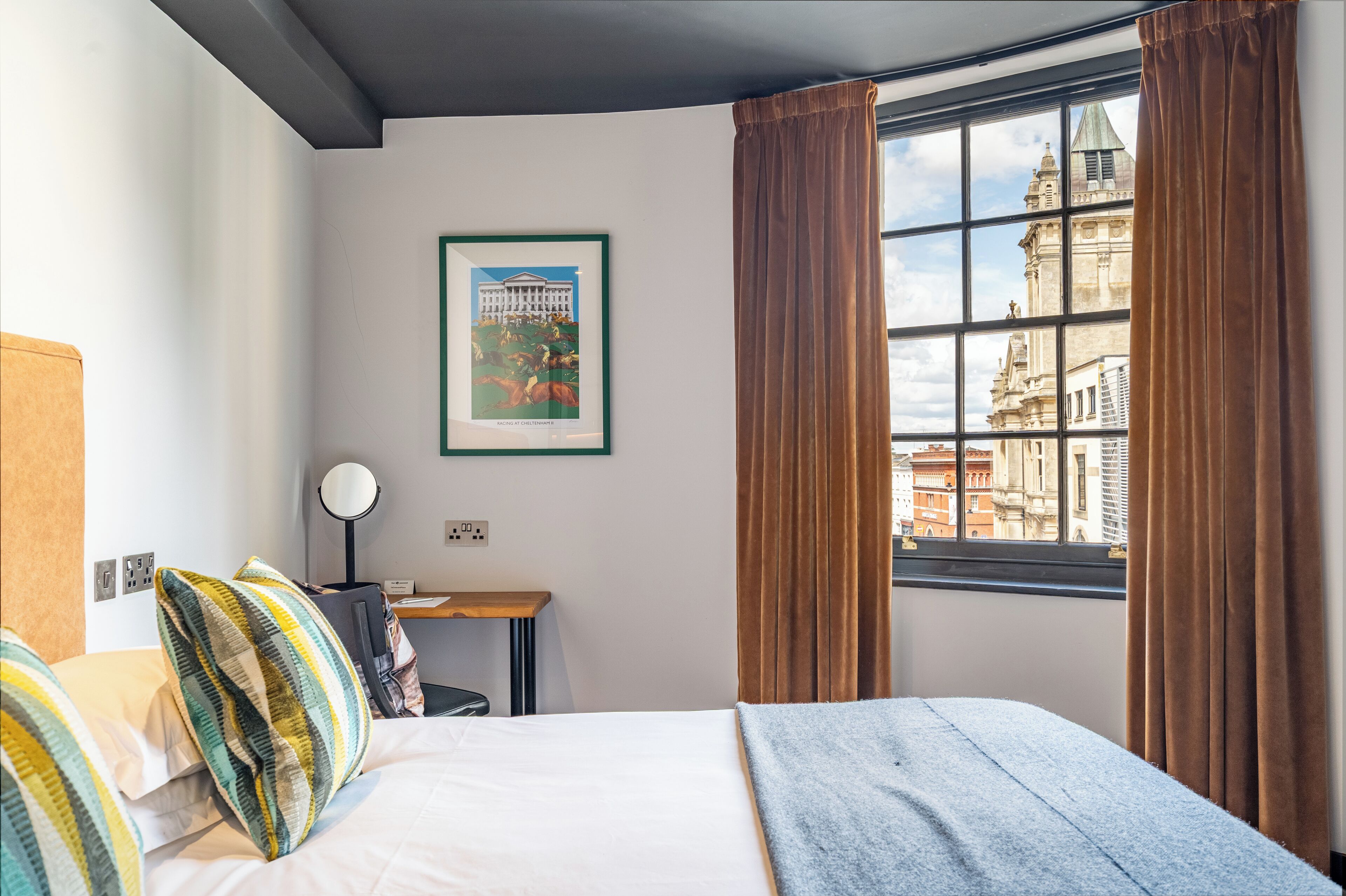 Habitació d'hotel amb un llit, escriptori, cadira, làmpada i finestra amb cortines.