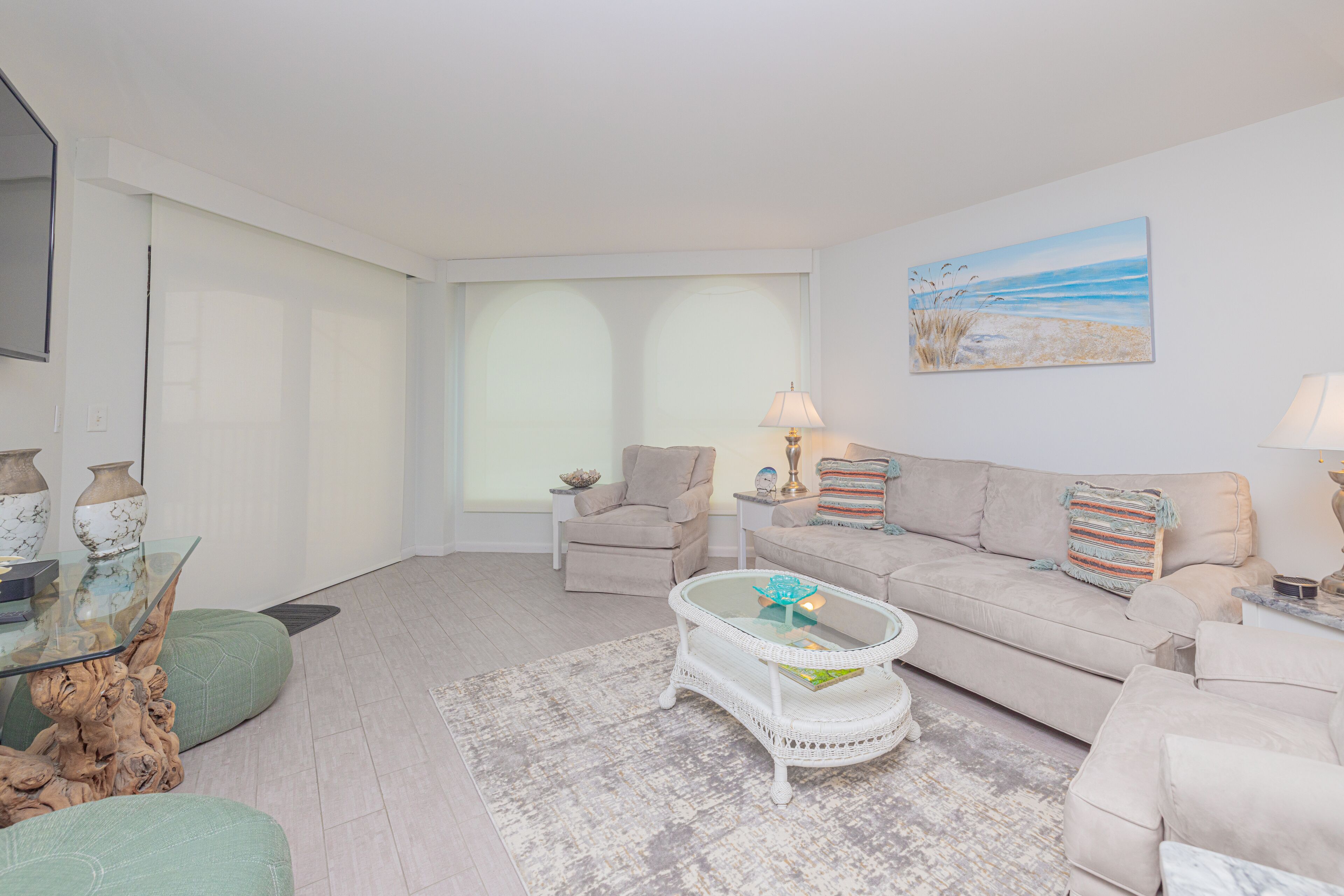 Una sala de estar con un sofá color beige, una mesa de centro de vidrio, un sillón color beige y un cuadro de una playa en la pared.