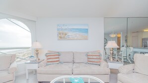 A living room with a beige sofa, two side tables with lamps, a glass coffee table, and a painting of a beach scene. - Oceanfront Condo in Wild Dunes! Community Pool! Great View Looking at Ocean! (Isle Of Palms)