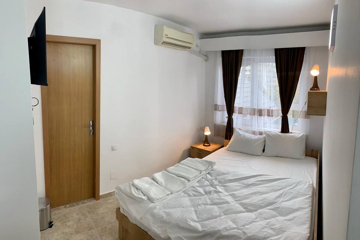 A bedroom with a bed, a nightstand, an air conditioning unit, a door, and a window with curtains.