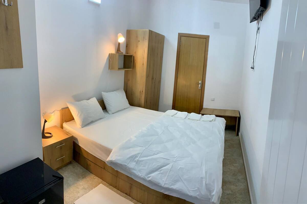A bedroom with a wooden bed frame, white bedding, a bedside lamp, a wooden wardrobe, and a small bedside table.