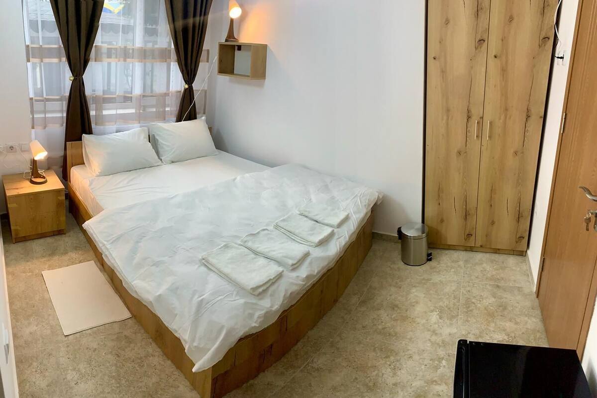 A neatly made bed with white linens, a bedside lamp, a wooden wardrobe, and a small trash bin.