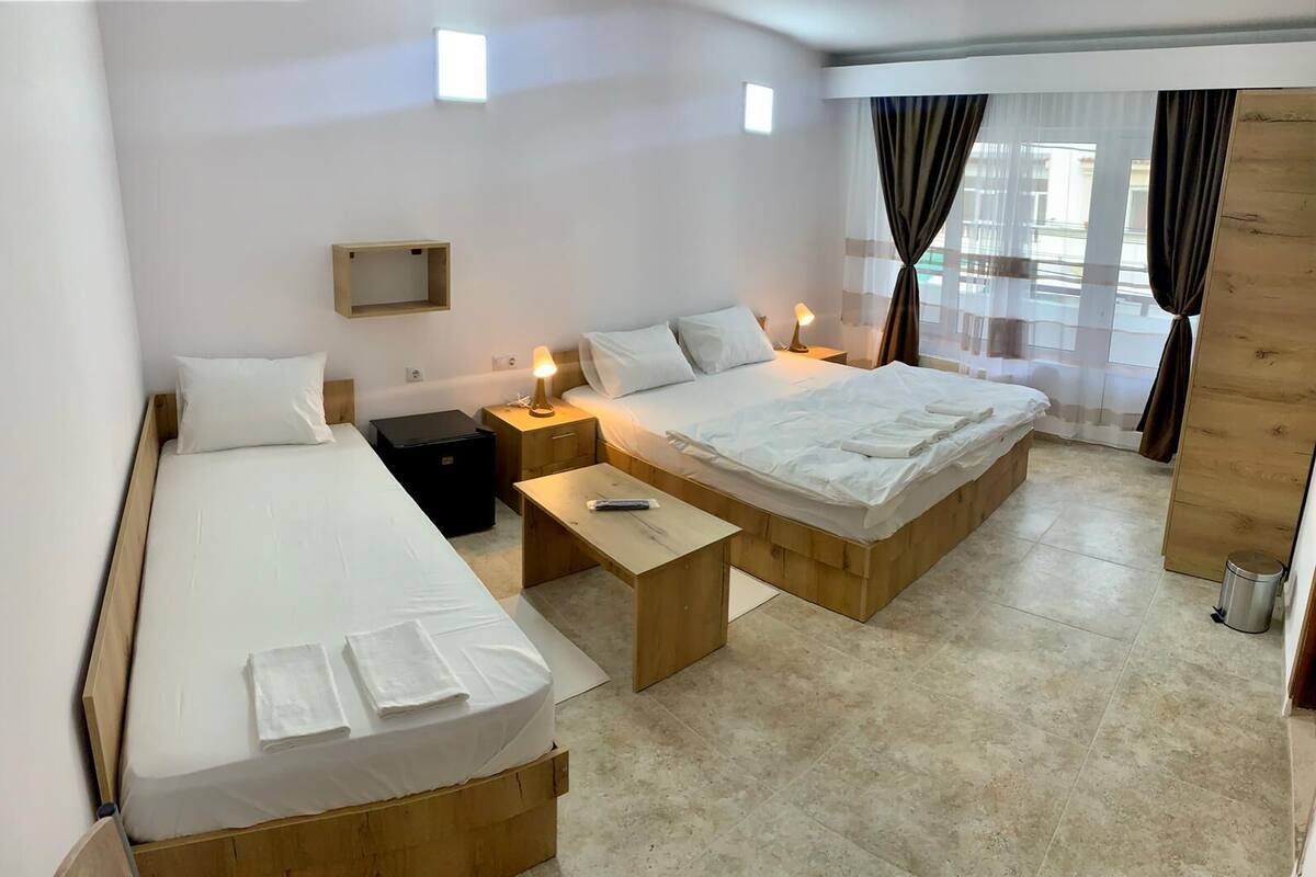 A hotel room with two beds, a small table, bedside lamps, a wooden cabinet, and a window with curtains.
