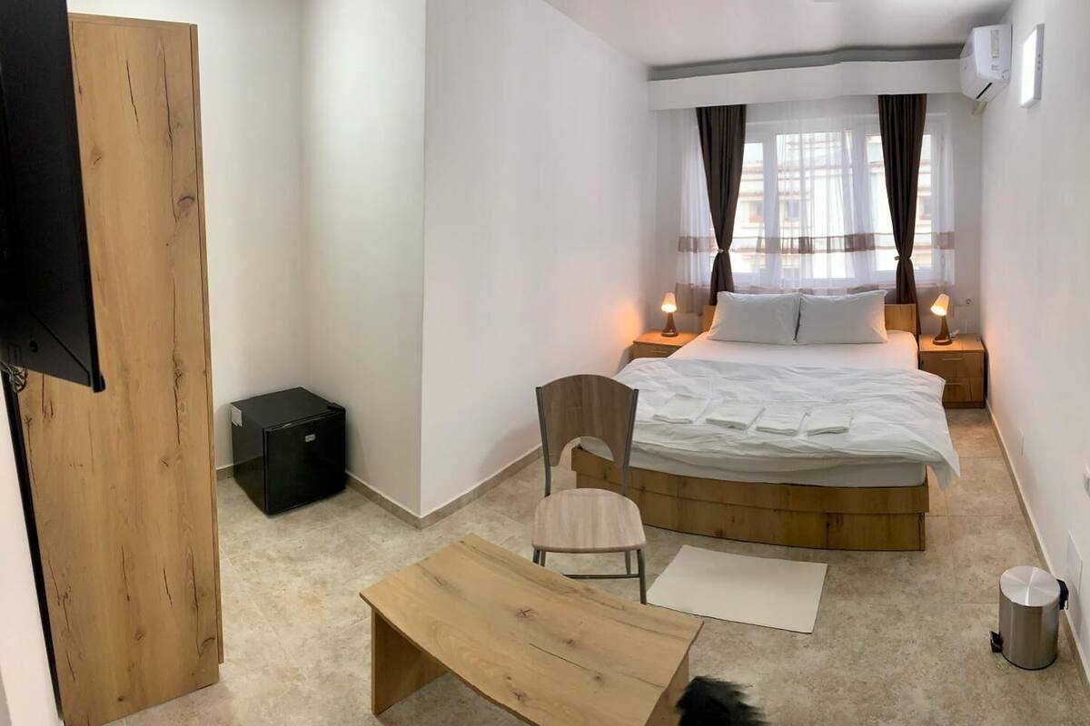 A compact hotel room with a bed, bedside tables, a chair, a small table, and a trash bin.