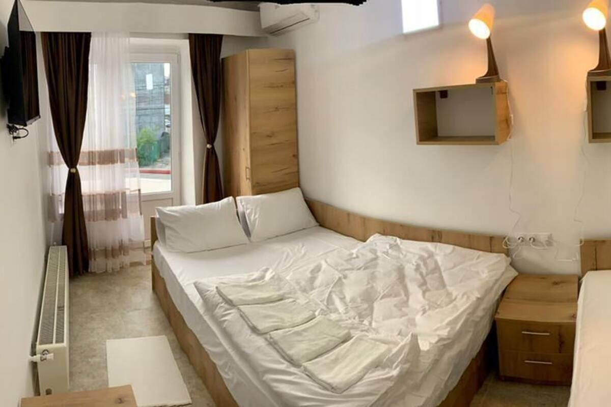 A bedroom with two beds, a wooden wardrobe, a small bedside table, and a window with curtains.