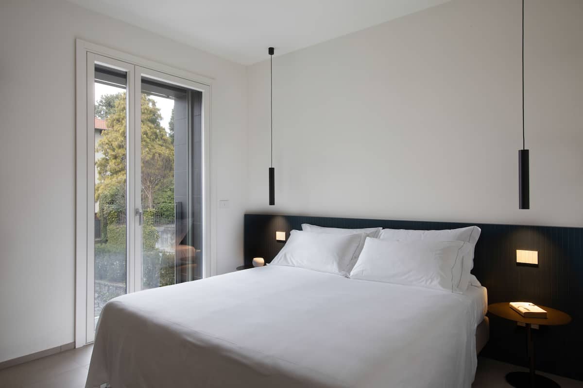 A modern hotel room with a large bed, a glass door leading to a balcony, and two pendant lights.