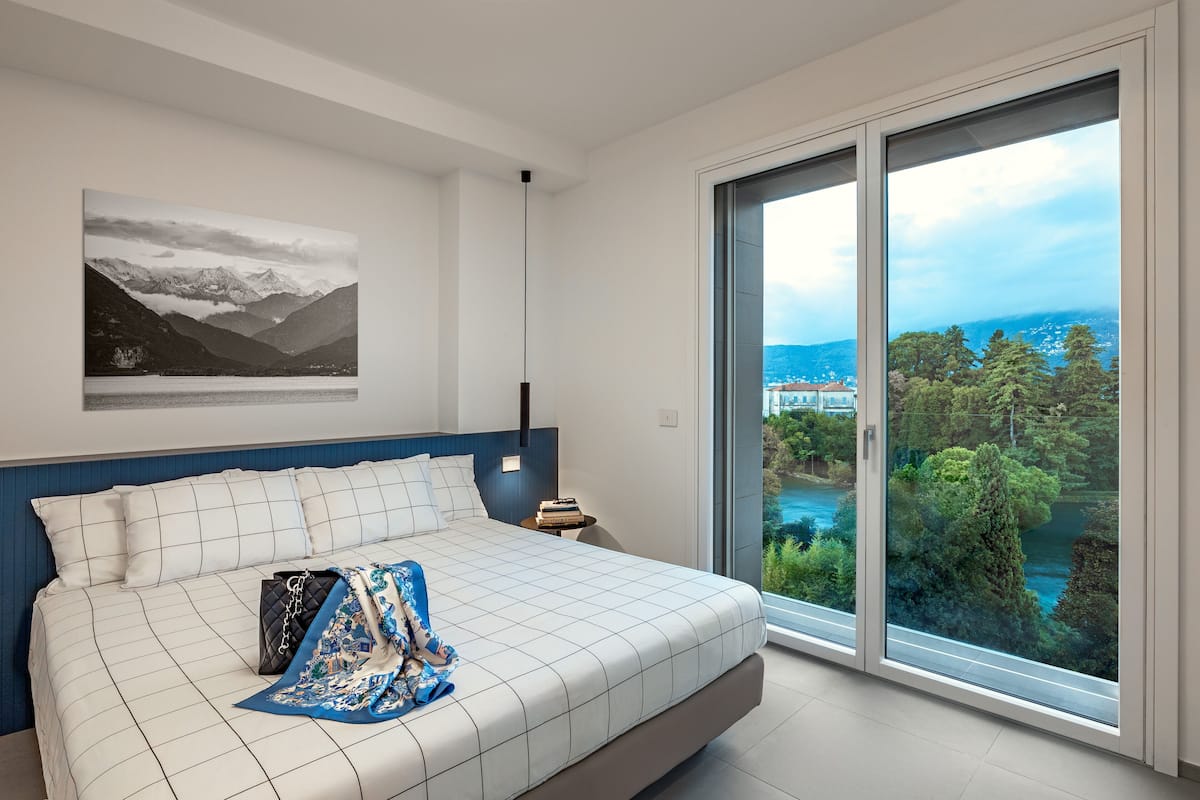 A bedroom with a bed, a nightstand, a large window with a view of a lake and mountains, and a painting on the wall.