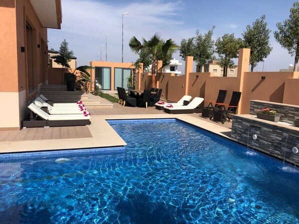 Une piscine entourée d’un petit muret et de palmiers, avec des chaises longues et une petite table à proximité.