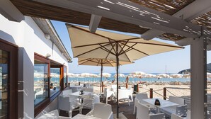 An outdoor seating area with white tables and chairs, a large umbrella, and a view of the beach and sea. - Hotel Delfino (Marciana)