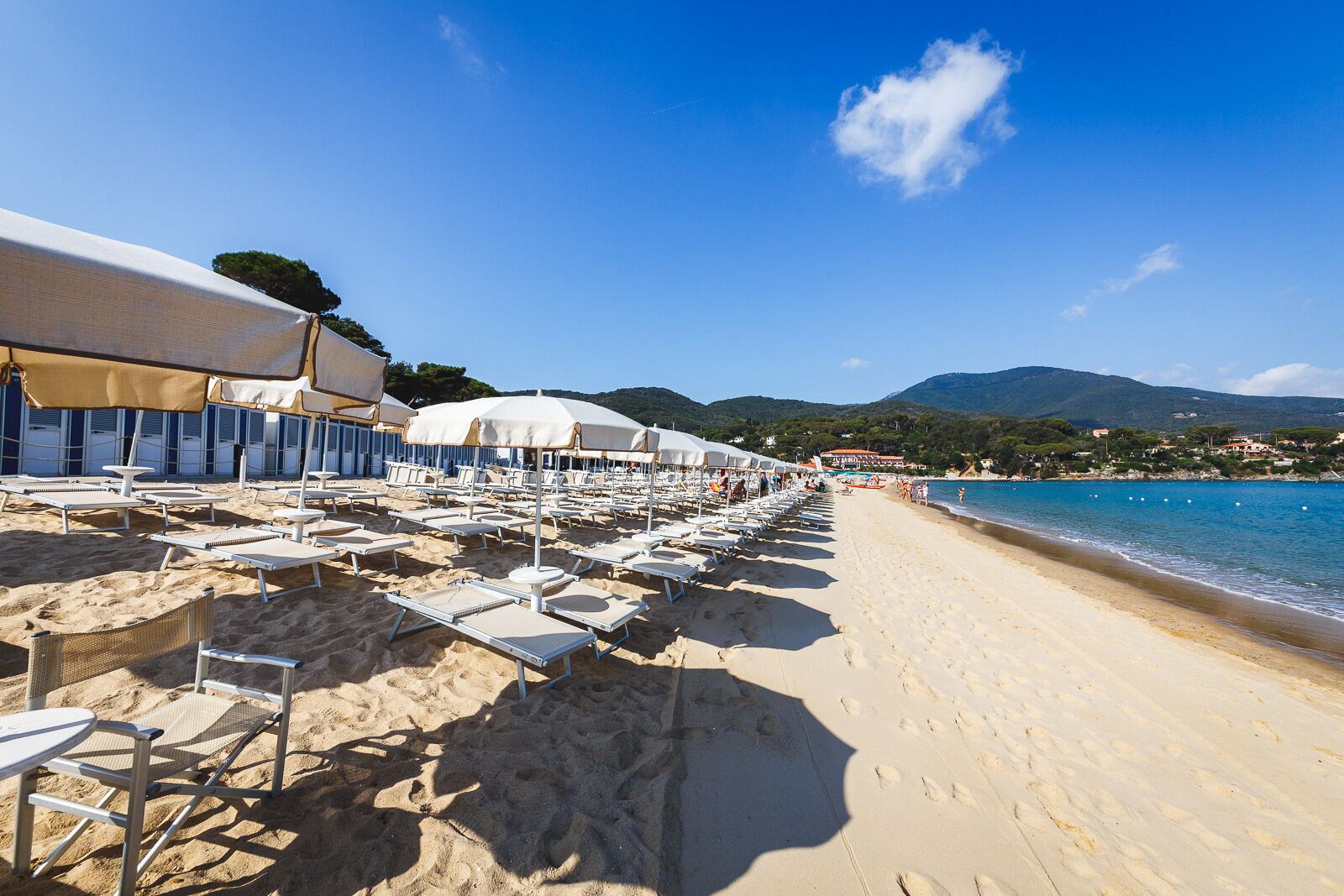 Une plage bordée de rangées de transats et de parasols, un ciel bleu azur et une vue sur la mer et les collines lointaines.