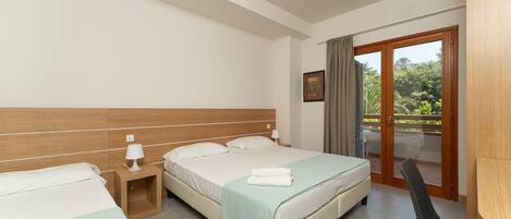 A hotel room with two beds, a wooden headboard, a nightstand with a lamp, a chair, and a balcony with a view of greenery.