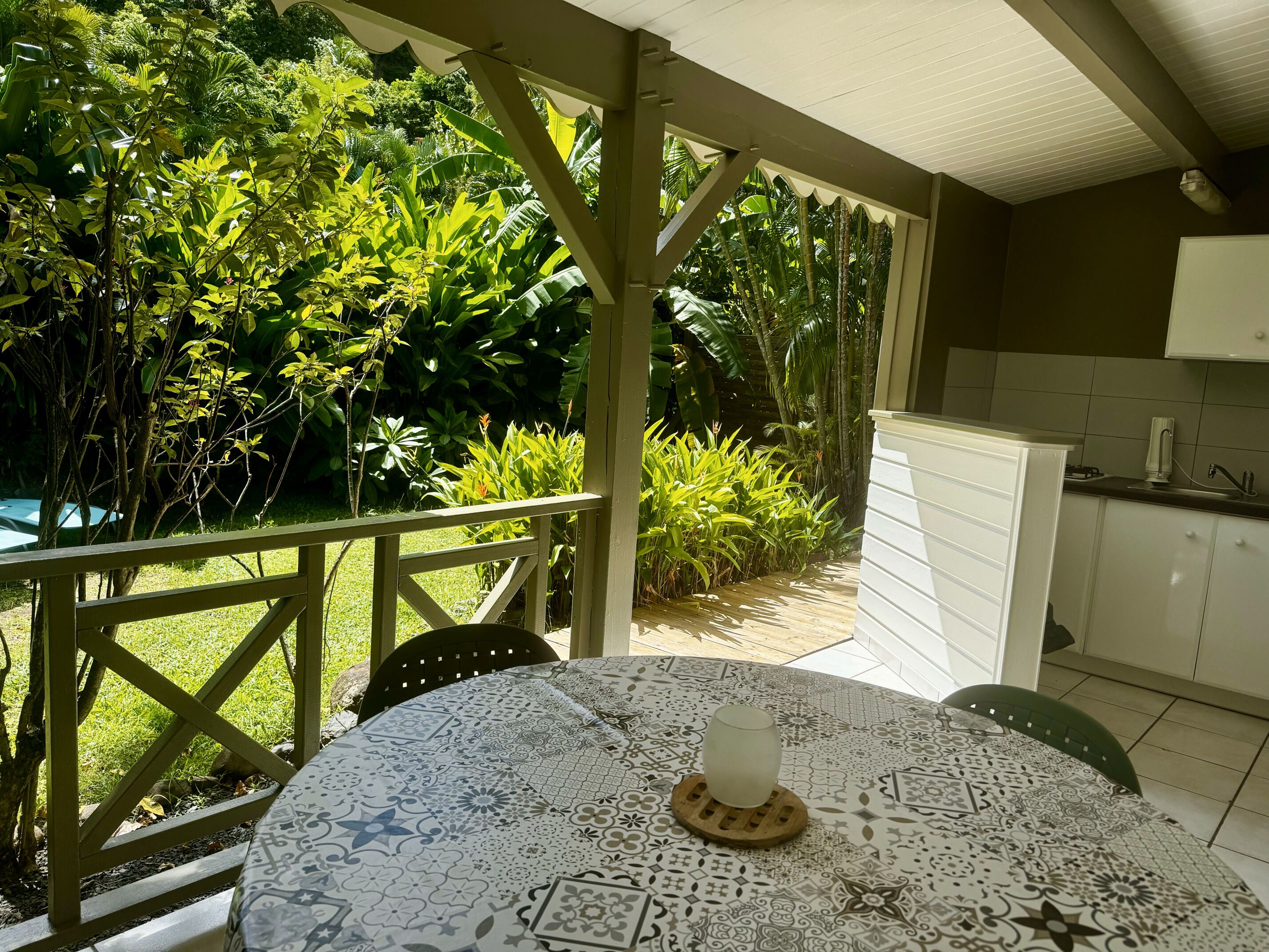 En overdækket terrasse med et bord og stole, omgivet af frodig vegetation.