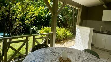 En overdækket terrasse med et bord og stole, omgivet af frodig vegetation.