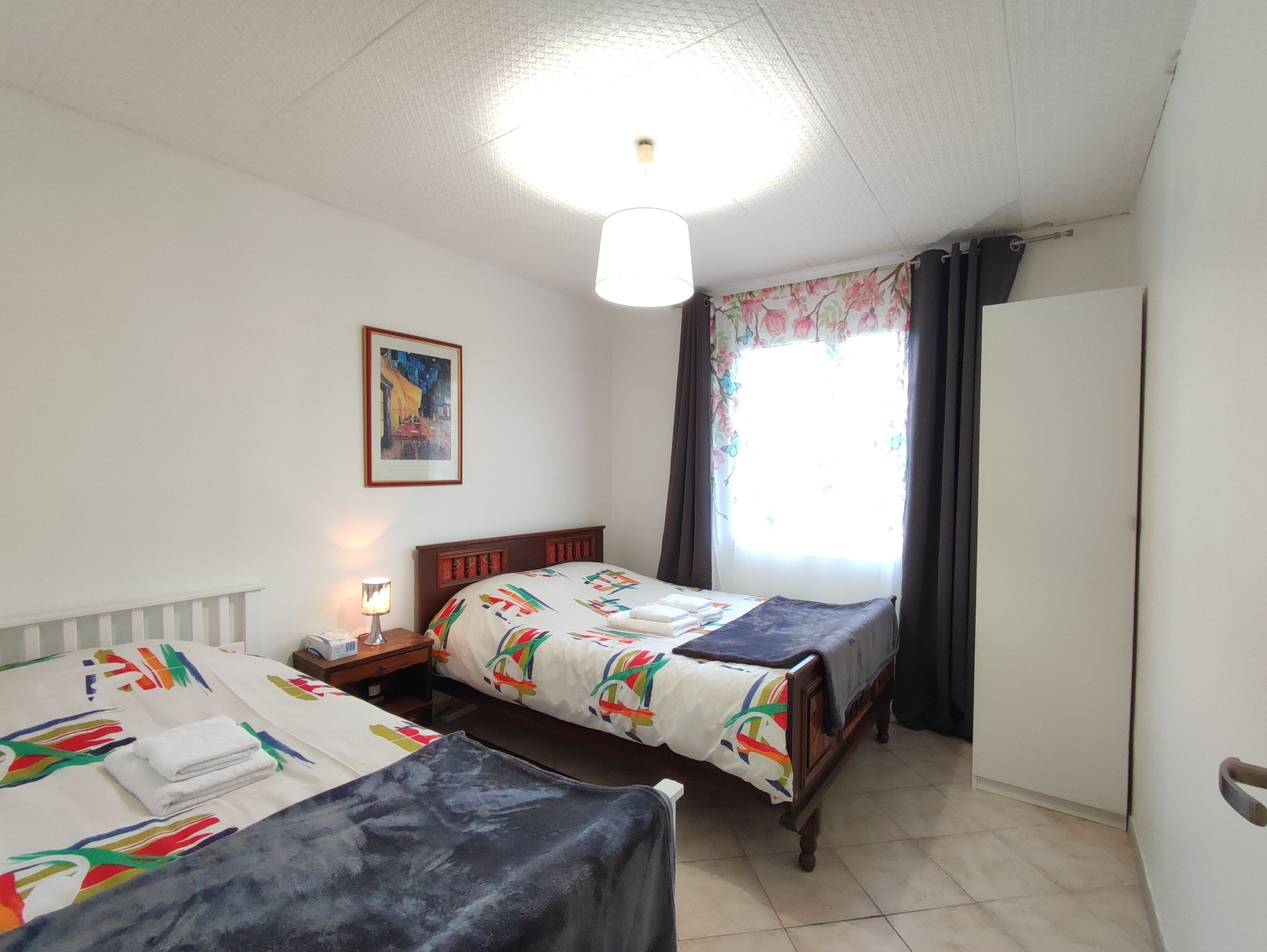 A bedroom with two beds, a nightstand, a bedside lamp, a framed picture on the wall, and a window with curtains.